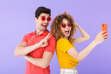 Portrait of fashion couple smiling and showing peace sign while taking selfie photo on cellphone in trendy sunglasses