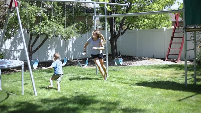Slow Motion shot of a girl playfully chasing her younger brother around in the backyard by the playground.