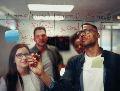 Successful Young Businessmen Writing On Transparent Glass Board With Red Marker In Office Meeting