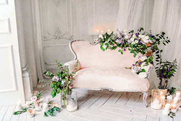 vintage sofa of soft pink color, decorated with flowers and greens, stands in a classic room on a white wooden floor surrounded by lighted candles in glass candlesticks near  large window and curtains