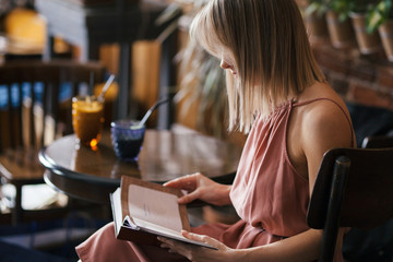 beautiful pensive calm relaxed girl blonde in ash rose color dress sits at a table in a retro vintage style cafe with a mug of drink on table and reading a book