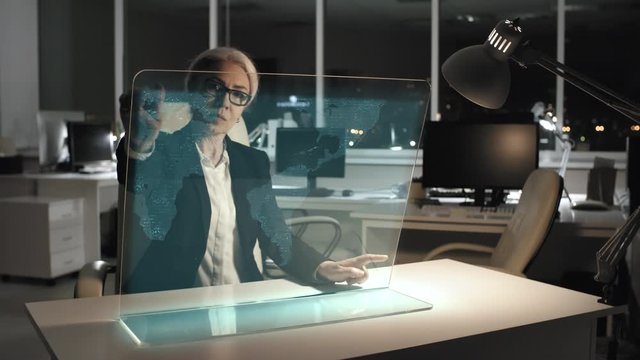 middle aged professional businesswoman tapping on AR computer screen and analyzing business data while working late in dark office