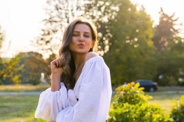 Young beautiful woman with bare shoulders in white blows a kiss in the park in summer at the sunset