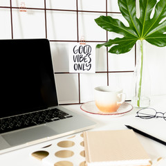 White desk with mood board, laptop, stationery, mug of coffee and green tropical leaf