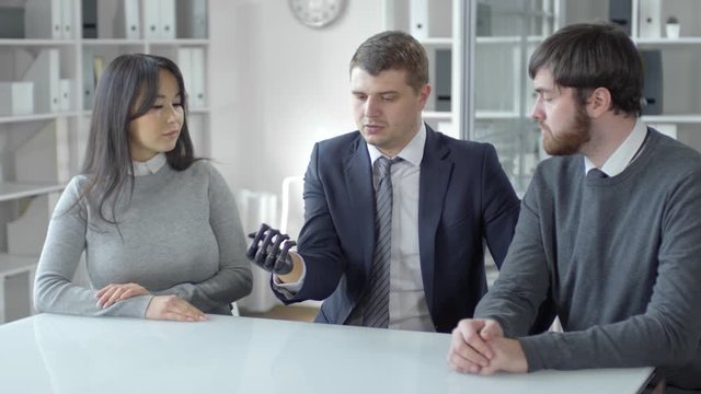 Businessman with prosthetic hand holding holographic globe projected in the air and discussing it with male and female colleagues while working at desk in the office