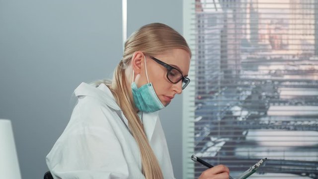 Profile view of female scientist in eyeglasses and lab coat writing notes and then drawing attention on something else in front. She putting on protective mask. Woman is very surprised.