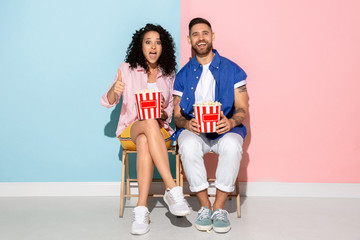 Young emotional caucasian couple in bright casual clothes posing on pink and blue background. Concept of human emotions, facial expession, relations, ad. Man and woman watch cinema with popcorn.