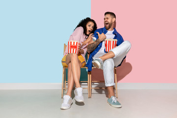 Young emotional caucasian couple in casual clothes posing on pink and blue background. Concept of human emotions, facial expession, relations, ad. Man and woman watch cinema with popcorn, look scared.