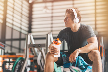Portait of athletic man in headphones looking aside while listening to music and holding a towel and a classic fitness shaker with pre-workout drink in it. Horizontal shot - obrazy, fototapety, plakaty
