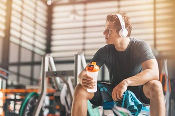Foto op Canvas Portait of athletic man in headphones looking aside while listening to music and holding a towel and a classic fitness shaker with pre-workout drink in it. Horizontal shot © Adamov
