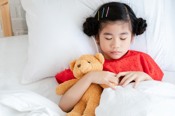 Asian kid girl wearing a red shirt hugs Teddy bear on the white bed. Have a flu Causing weak health to be unhealthy Must get enough rest Or go to the hospital for a specialist medical consultation