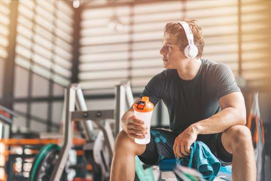 Portait of athletic man in headphones looking aside while listening to music and holding a towel and a classic fitness shaker with pre-workout drink in it. Horizontal shot