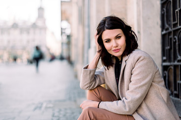 Obraz premium Close-up portrait of pretty girl with short brunette hair at street background wearing beige coat, make up. Smiling and sensual young woman posing while sitting on stairs