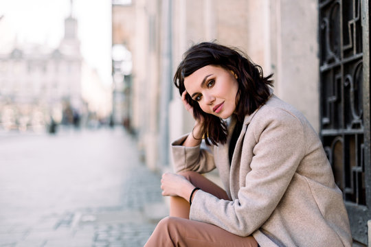 Close-up portrait of pretty girl with short brunette hair at street background wearing beige coat, make up. Smiling and sensual young woman posing while sitting on stairs