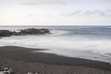 Fototapeta na wymiar Mesa del mar volcanic beach sand and rocks, long exposure photography, with Atlantic ocean waves, horizon with sunset light, Tacoronte, Tenerife, Canary islands, Spain