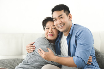 Asian mother hugging her son and sitting on sofa at home, lifestyle concept.