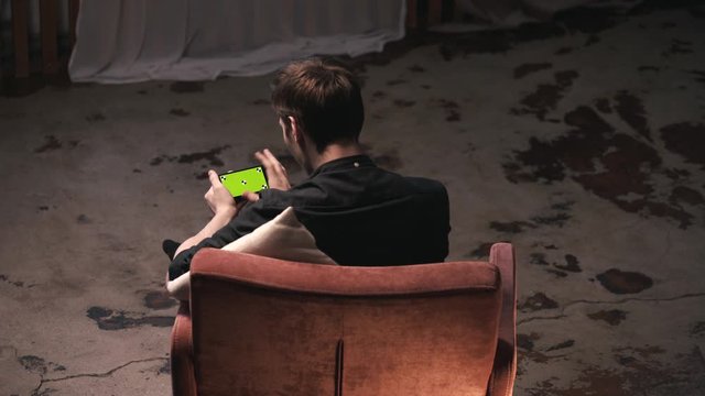 Attractive young man with brown hair sitting in comfortable chair with white pillow and sliding on iPhone green screen in dark room. Stock footage. Chroma key