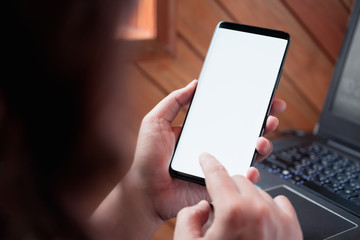 Close up image of smart phone mockup, business woman holding and touch smartphone with white blank screen template with blur laptop background.