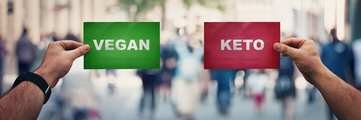 People hands holding two papers over a crowded street background comparing Vegan vs Ketogenic...