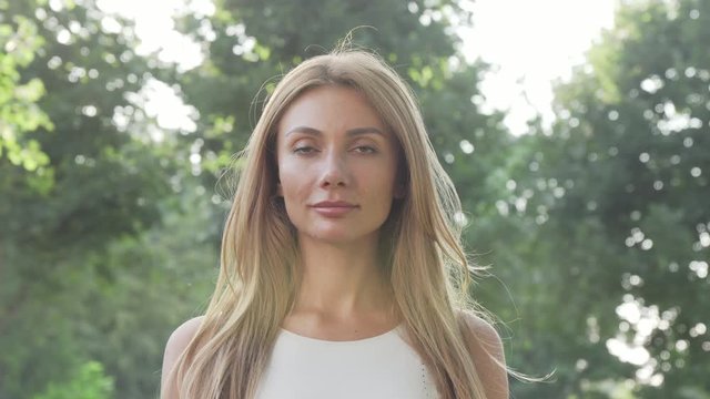 Beautiful happy woman enjoying relaxing outdoors in the park in summer evening. Gorgeous female breathing fresh air outdoors, smiling to the camera. Happiness concept. 4k footage