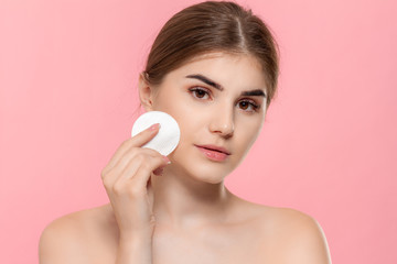 Beautiful model applyes cosmetic product to skin with white cotton sponge. Model looks at the camera isolated over pink background. Concept of beauty and health treatment.