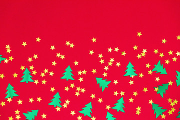 Green metallic foil christmas trees and gold stars confetti sparse on red background. Simple holiday concept. Design template. Frame with copy space. Winter festive backdrop. Top view, flat lay.