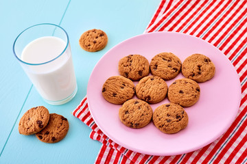 A glass full of milk, chocolate chip cookies in a pink plate and a red tablecloth on blue wooden table. Close up view from top.
