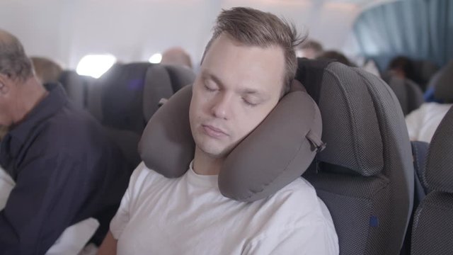 Young man sitting on an airplane, wearing a neck pillow while sleeping