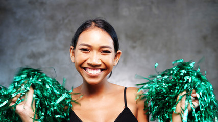 Portrait of beautiful young smiling girl with green cheerleader pom-poms over concrete wall...
