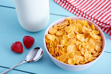 Bowl of corn flakes cereal, strawberries, a bottle of milk, napkin and spoon on blue wooden country style table. View from above.