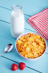 Bowl of corn flakes cereal, strawberries, a bottle of milk, napkin and spoon on blue wooden country style table. View from above.