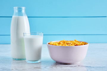 Bottle of milk, bowl of cereal corn flakes and a glass full of milk on a wooden table against blue wooden background. Close up view.