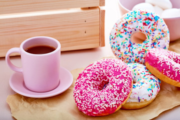 Colorful donuts on paper with pink coffee cup, marshmallows and a crate on table. Close up view.