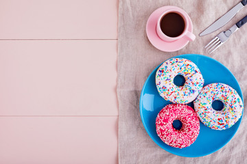 Colorful donuts in a plate with cappuccino coffee cup, fork and knife on a tablecloth over pink wooden table background with copy space. Top view.