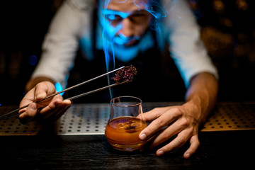 Professional bartender adding chilled melting caramel with twezzers to the cocktail with ice cubes under blue light
