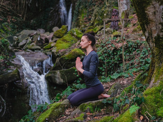 Young slim woman practicing yoga outdoors in moss forest on background of waterfall. Unity with nature concept. Girl meditates sitting