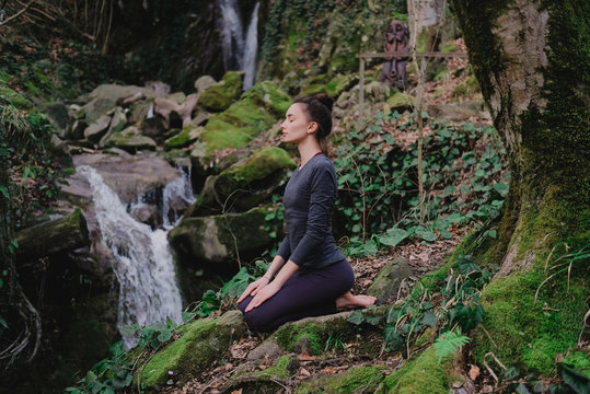 Young slim woman practicing yoga outdoors in moss forest on background of waterfall. Unity with nature concept. Girl meditates sitting
