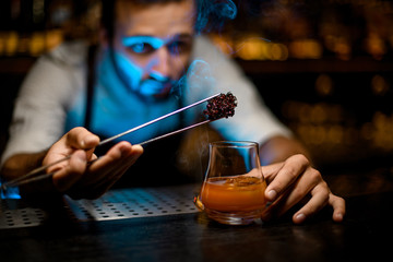 Bartender adding chilled melting caramel with twezzers to the cocktail with ice cubes under blue light