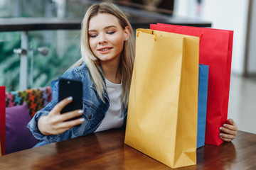Blonde woman in a jeans jacket with shopping bags sitting in cafe at shopping mall. She is taking...