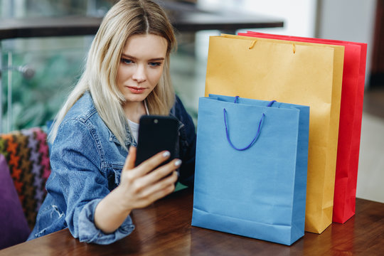 Beautiful blonde woman in a jeans jacket with shopping bags sitting in cafe at shopping mall. She is taking pictures of herself on her phone doing a sephi. Concept relaxing after shopping
