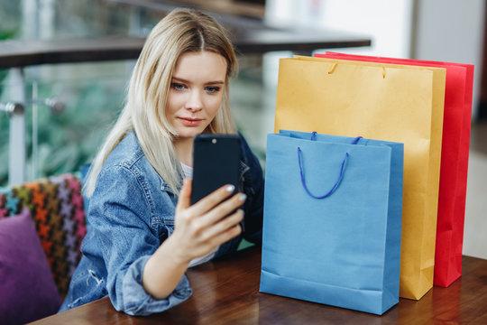 Woman in a jeans jacket with shopping bags sitting in cafe at shopping mall. She is taking pictures of herself on her phone doing a sephi. Shopping bags on the table. Concept relaxing after shopping