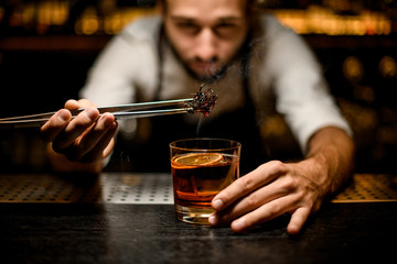 Male bartender adding chilled brown caramel with twezzers to the cocktail with ice cubes