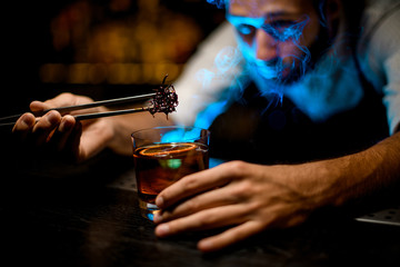 Male bartender adding chilled melting caramel with twezzers to the cocktail with dried orange under blue light