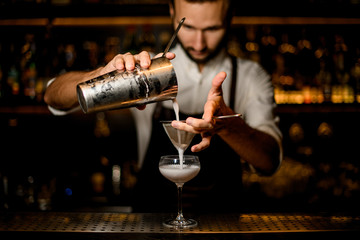 Professional bartender pouring a white cocktail from the steel shaker to the glass through the sieve