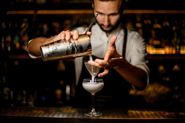Male bartender pouring a white cocktail from the steel shaker to the glass through the sieve