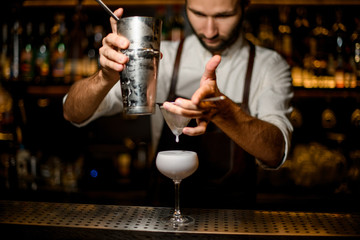 Bartender pouring a white cocktail from the steel shaker to the glass through the sieve