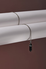 Close-up shot of a steely chainlet with a pendant in the form of screw with a fitted-in dark gemstone similar to a black onyx. The glazy gemstone is elongated and conical.  © RedUmbrella&Donkey
