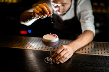 Professional bartender sprinkling on the scum of the light violet cocktail with a yellow and purple flower petals