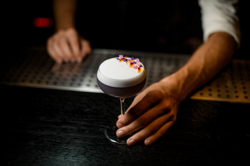 Professional bartender serving a cocktail with white scum decorated with flower petals on the bar counter