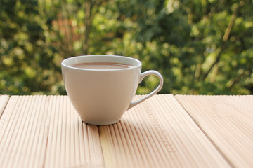 white cup with coffee on a light wooden table against a green summer garden, close-up, copy space